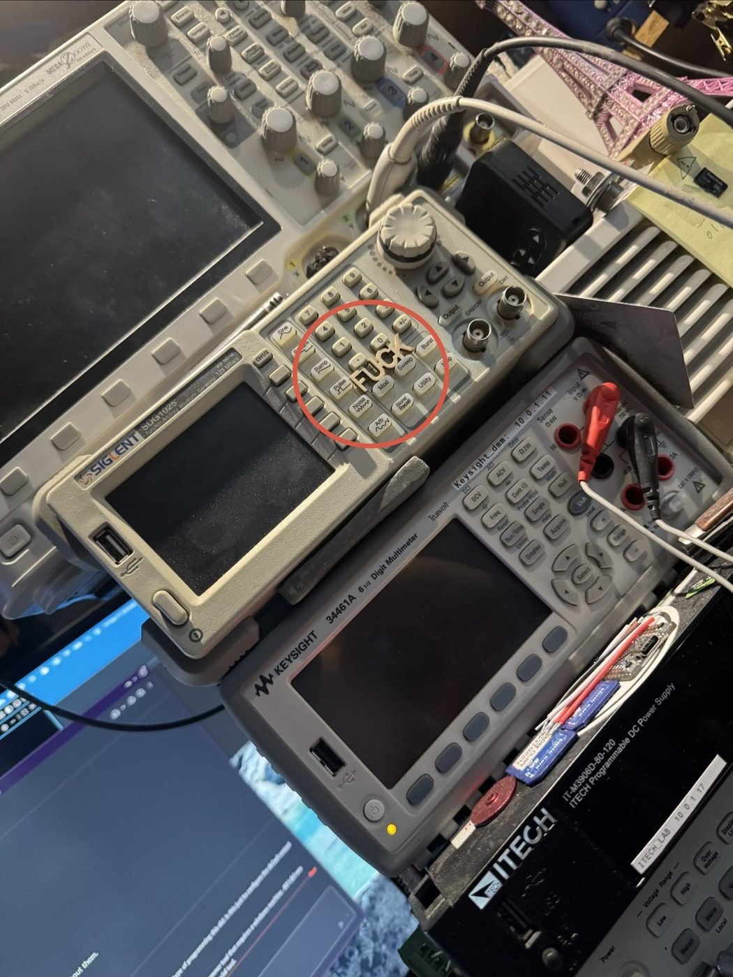 A small wooden FUCK resting on the front panel of a Siglent function generator on a crowded electronics bench, circled in red